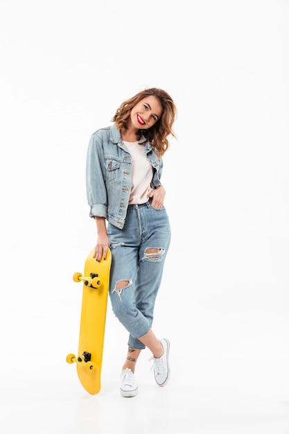 Longitud total mujer alegre en ropa de mezclilla posando con patineta sobre pared blanca