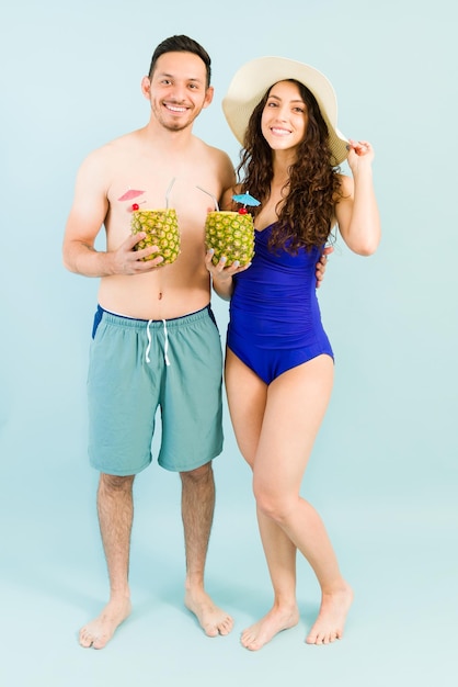 Longitud total de un novio y una novia hispanos bebiendo un cóctel de piña colada junto a la piscina. Atractiva pareja yendo de vacaciones de verano