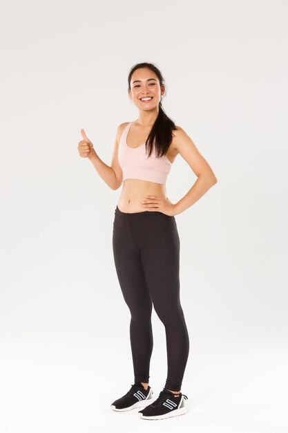 Longitud total de sonriente satisfecha, linda chica asiática de fitness, deportista en ropa activa mostrando el pulgar hacia arriba y sonriendo complacida, orgullosa athelte femenino ganar objetivo de entrenamiento diario, fondo blanco.