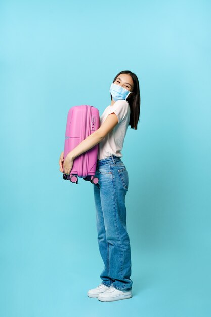 Longitud total de turista mujer emocionada abrazando la maleta y sonriendo con mascarilla médica de cov ...