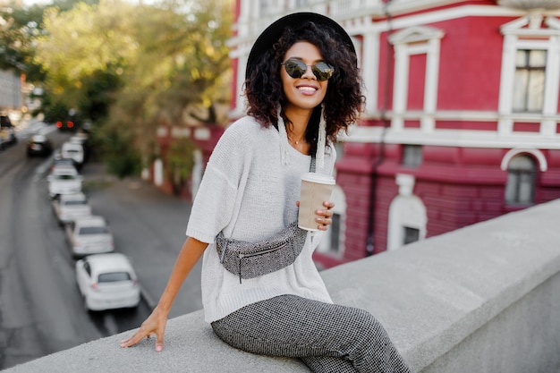 Foto gratuita look de moda callejera. elegante chica negra sentada en el puente y sosteniendo una taza de café o té durante su tiempo libre. mujer independiente. llevaba sombrero negro y gafas de sol.