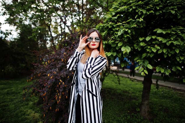 Look de mujer de moda con pantalones de cuero de chaqueta de traje a rayas blancas y negras y gafas de sol posando contra arbustos en la calle Concepto de chica de moda