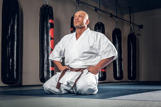 Foto gratuita el luchador de karate con la cabeza rapada se sienta en el tatami en un gimnasio.