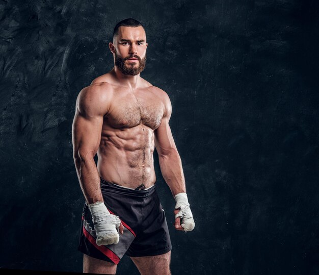 Un luchador musculoso y guapo con el torso desnudo está demostrando su poder en un estudio fotográfico oscuro.