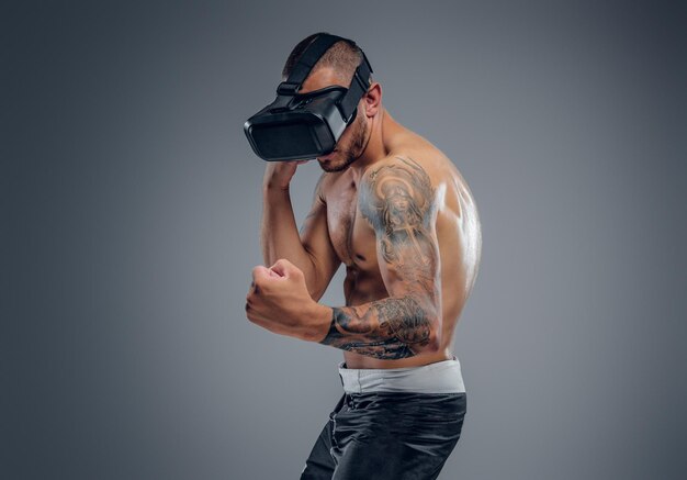 Un luchador tatuado sin camisa con gafas de realidad virtual en la cabeza aislado en un fondo gris.