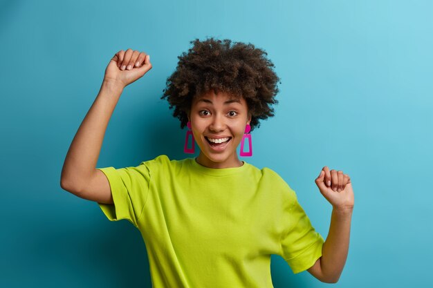 Lucky complacida joven de pelo rizado baila con las manos en alto, se divierte y expresa emociones positivas, libertad y felicidad, se siente como campeona, vestida con una camiseta verde brillante, ganó su objetivo