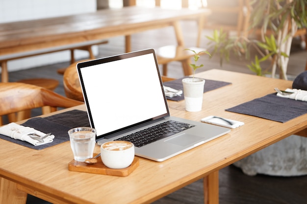 Lugar de trabajo de un autónomo desconocido cuando no hay nadie cerca: toma minimalista de una taza de café, un vaso de agua, un teléfono celular y una computadora portátil genérica