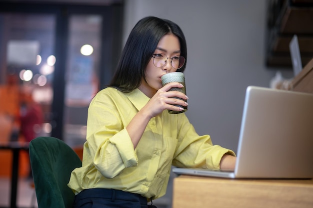 Lugar de trabajo. Linda mujer joven asiática en anteojos en la computadora portátil tomando café