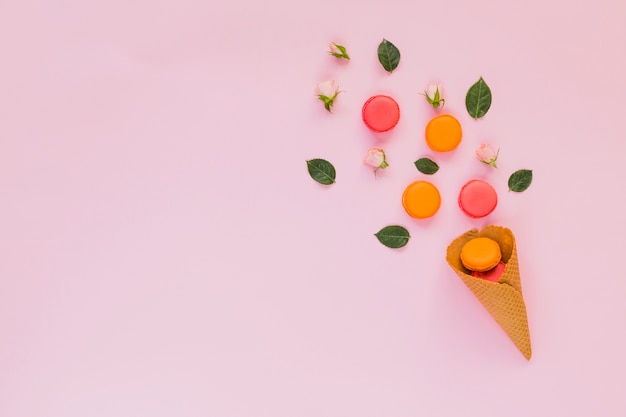 Macarrones de colores; Rosa; y hojas dispuestas sobre el cono de waffle sobre fondo rosa