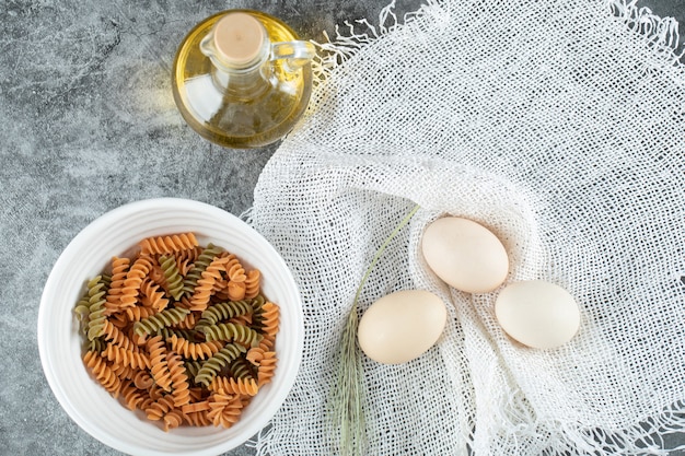 Macarrones en espiral sin preparar en un plato blanco con tres huevos y una botella de aceite