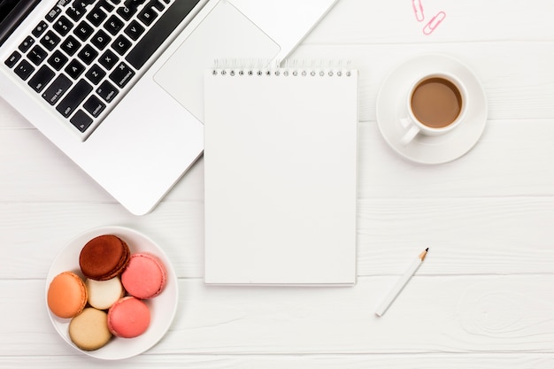 Macarrones y taza de café coloridos con la libreta espiral en el ordenador portátil sobre el escritorio de oficina de madera