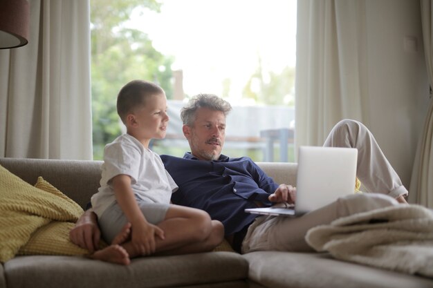 Macho adulto acostado en el sofá con su hijo y usando la computadora portátil bajo las luces