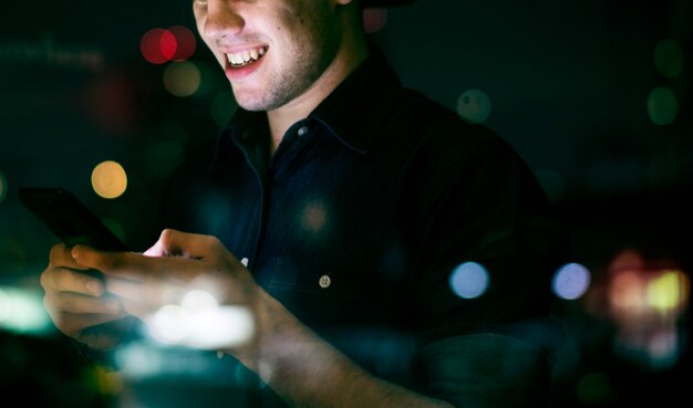 Macho adulto joven feliz usando un teléfono inteligente en un paisaje urbano de noche