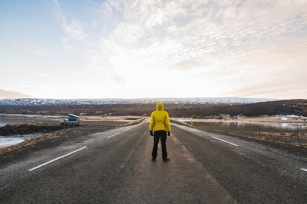 Macho con una chaqueta amarilla de pie en la carretera rodeada de colinas cubiertas de nieve en Islandia