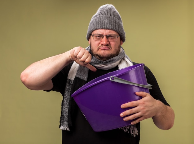 Macho enfermo de mediana edad disgustado con gorro y bufanda de invierno sosteniendo un cubo de plástico mostrando el pulgar hacia abajo aislado en la pared verde oliva