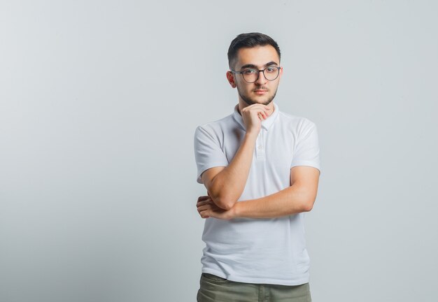 Macho joven apoyando la barbilla en la mano en camiseta blanca, pantalones y mirando pensativo