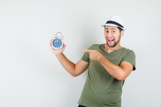 Macho joven apuntando al despertador en camiseta verde y sombrero y mirando contento