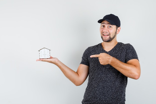 Macho joven apuntando al modelo de la casa en camiseta y gorra y mirando alegre