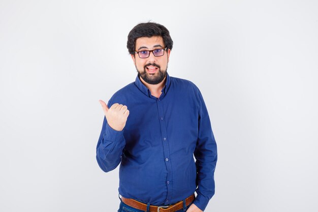 Macho joven apuntando a un lado en camisa azul real, gafas, vista frontal.