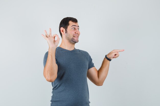 Macho joven apuntando a un lado mostrando gesto ok en camiseta gris y mirando alegre