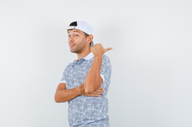 Macho joven apuntando con el pulgar hacia fuera en camiseta y gorra y mirando astuto