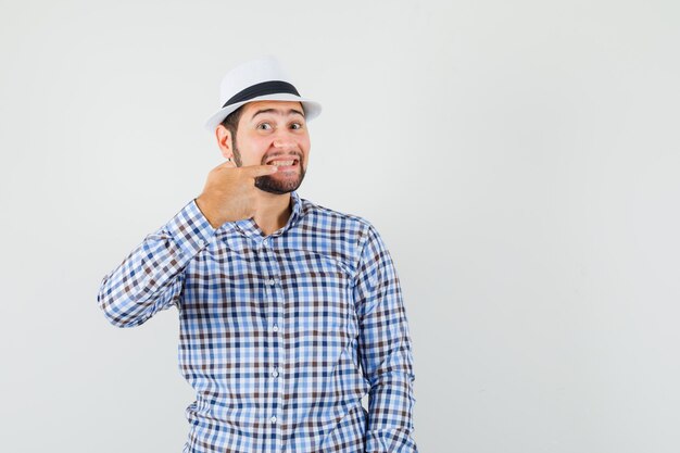 Macho joven apuntando a sus dientes en camisa a cuadros, sombrero y mirando alegre