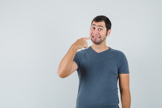 Macho joven apuntando a sus dientes en camiseta gris y mirando alegre