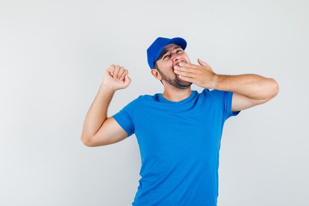 Macho joven bostezando y estirándose en camiseta azul y gorra y mirando soñoliento