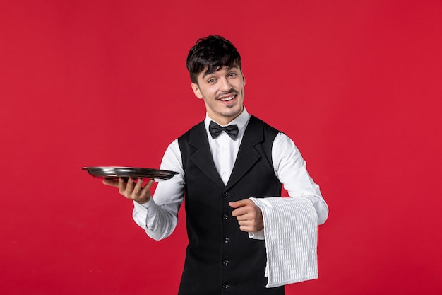 Macho joven camarero en uniforme con mariposa en el cuello y sosteniendo una bandeja de toalla sobre fondo rojo.
