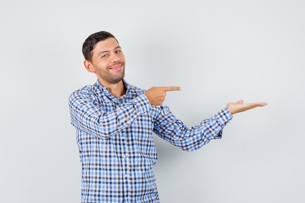 Macho joven en camisa a cuadros apuntando a su palma extendida a un lado y mirando alegre