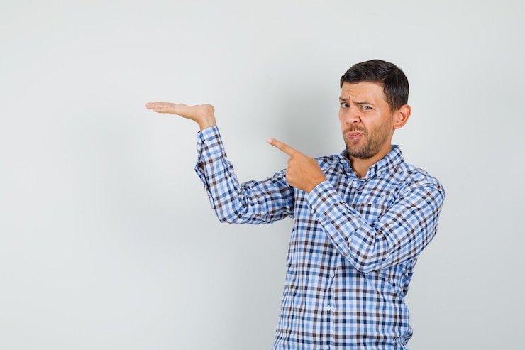 Foto gratuito macho joven en camisa a cuadros apuntando a su palma levantada y mirando vacilante