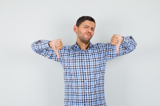 Macho joven en camisa a cuadros que muestra el doble pulgar hacia abajo y mirando disgustado