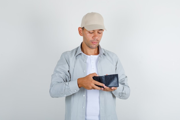 Macho joven en camisa con gorra mirando en billetera abierta y mirando con cuidado