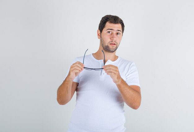 Macho joven en camiseta blanca mirando a cámara con gafas en mano