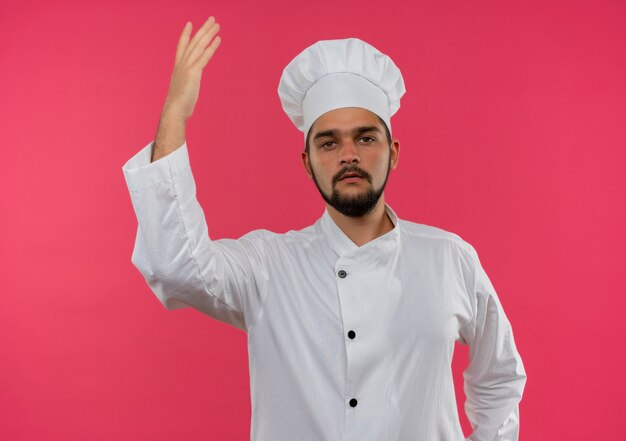 Macho joven cocinero en uniforme de chef levantando la mano y mirando