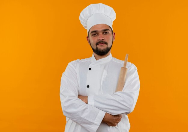 Macho joven cocinero en uniforme de chef de pie con postura cerrada y sosteniendo