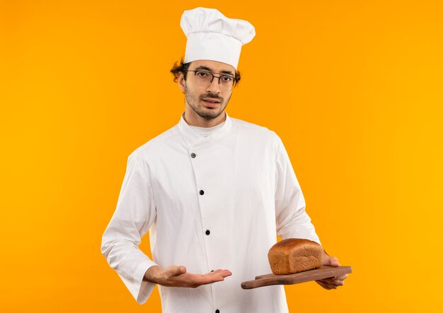 Macho joven cocinero vistiendo uniforme de chef y gafas sosteniendo y apunta con la mano al pan en la tabla de cortar