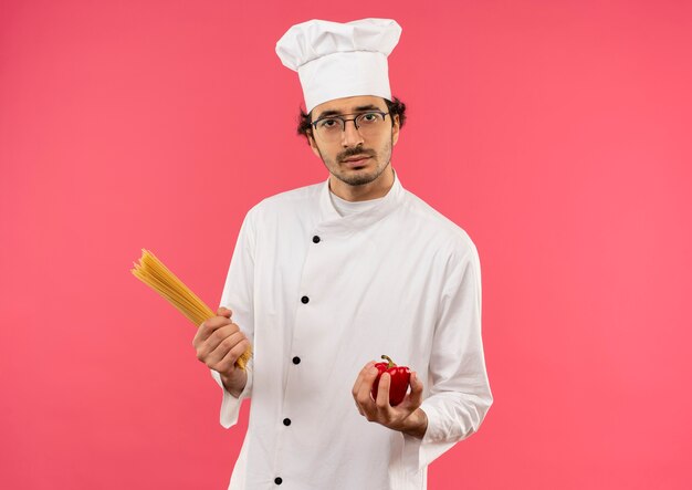 Macho joven cocinero vistiendo uniforme de chef y gafas sosteniendo espaguetis y pimienta