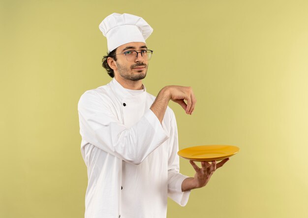 Macho joven cocinero vistiendo uniforme de chef y gafas sosteniendo la placa y fingiendo derramar sal