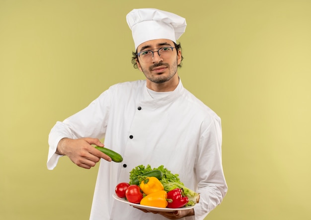 Macho joven cocinero vistiendo uniforme de chef y gafas sosteniendo verduras en la placa