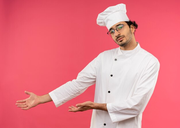 Macho joven cocinero vistiendo uniforme de chef y puntos de gafas con la mano al lado
