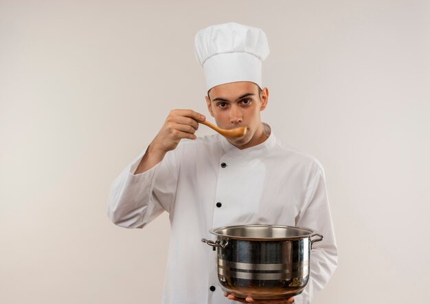 Macho joven cocinero vistiendo uniforme de chef sosteniendo una cacerola tratando de sopa de cuchara en la pared blanca aislada