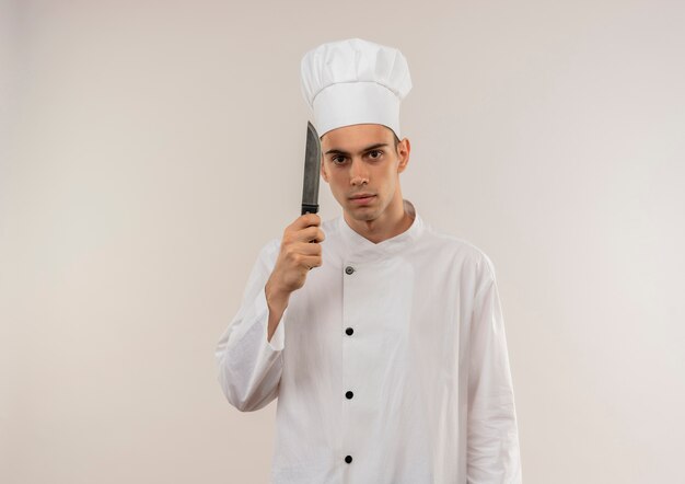 Macho joven cocinero vistiendo uniforme de chef sosteniendo un cuchillo en la pared blanca aislada con espacio de copia