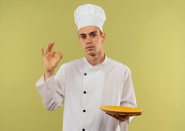 Macho joven cocinero vistiendo uniforme de chef sosteniendo la placa mostrando gesto okey en pared verde aislada con espacio de copia