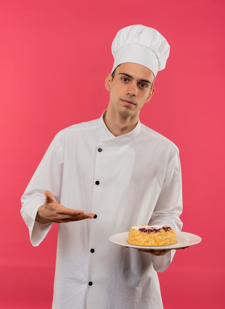 Macho joven cocinero vistiendo uniforme de chef sosteniendo y puntos con pastel de mano en la placa en la pared rosa aislada
