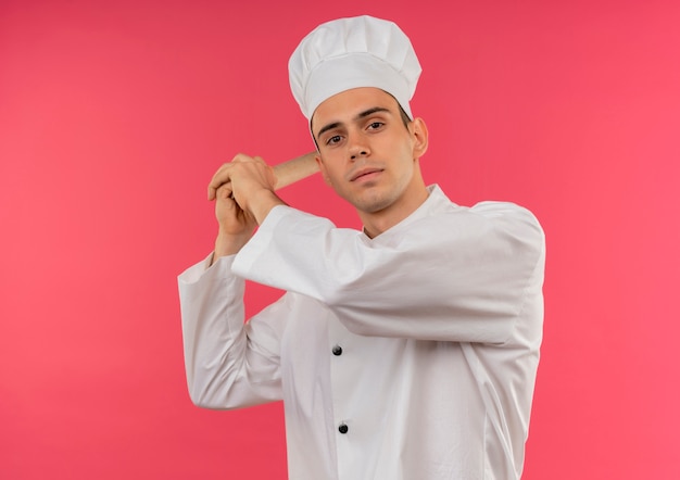 Macho joven cocinero vistiendo uniforme de chef sosteniendo rodillo alrededor del hombro en la pared rosa aislada