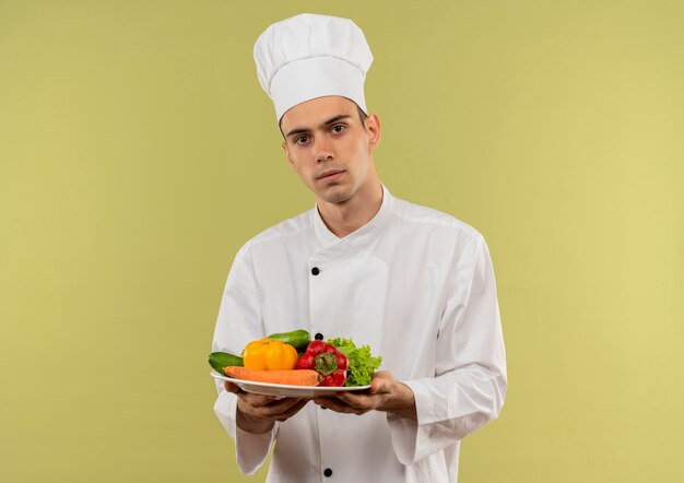 Macho joven cocinero vistiendo uniforme de chef sosteniendo verduras en la placa en la pared verde aislada con espacio de copia