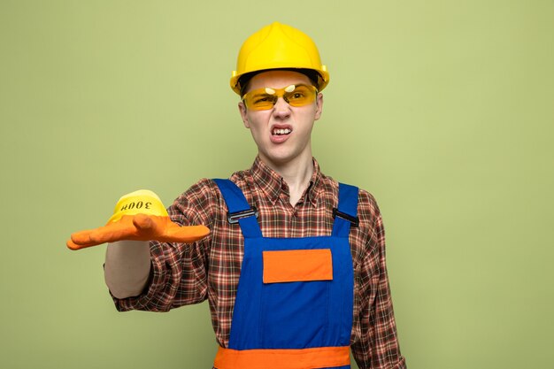 Macho joven constructor con uniforme y guantes con gafas aislado en la pared verde oliva
