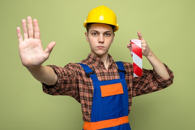 Macho joven constructor vistiendo uniforme con cinta adhesiva aislada en la pared verde oliva