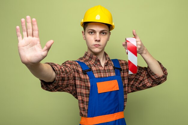 Macho joven constructor vistiendo uniforme con cinta adhesiva aislada en la pared verde oliva